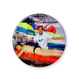 May include: A man wearing a white t-shirt with the word "Sandals" printed on it, is holding a drink and smiling at the camera. A woman is lying on a red towel on a wooden lounge chair. The man is standing behind the lounge chair. There is a green and yellow umbrella in the background.