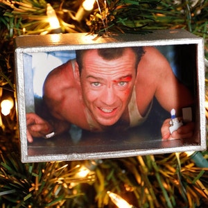 May include: A Christmas ornament featuring a framed image of a man with a bloody face, looking up at the viewer. The man is wearing a white tank top and is holding a lighter in his right hand.