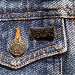 May include: Black enamel pin with gold lettering that reads "Michael Jackson".