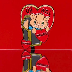 May include: A red heart-shaped pin with a cat inside holding a red heart that says "Whatever". The heart is surrounded by a gold frame and the words "Oh Well" are written on the top of the heart.