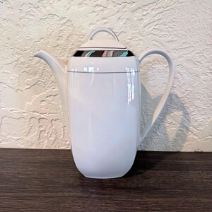 May include: White ceramic coffee pot with a black and colorful patterned band around the lid. The pot has a curved handle and a long spout. The design is simple and elegant, suitable for serving coffee or tea. The pot is placed on a dark wooden surface.