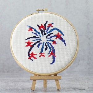 May include: A circular wooden embroidery hoop containing white Aida fabric with a cross-stitch design of a firework burst in red and dark blue. The hoop is mounted on a small, light-colored wooden display easel for upright presentation.