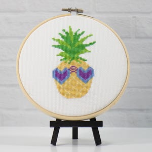 Puede incluir: Diseño de piña en punto de cruz en un bastidor de bordado de madera. La piña es amarilla con hojas verdes y gafas de sol moradas en forma de corazón. El bastidor se exhibe en un pequeño caballete negro.