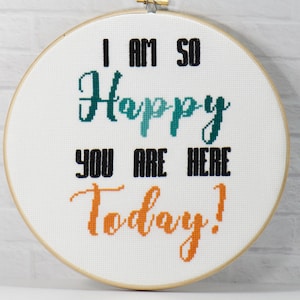 Può includere: Un telaio per punto croce con le parole "I AM SO Happy YOU ARE HERE Today!" in filo nero, verde acqua e arancione. Il testo è centrato su uno sfondo di tessuto bianco, incorniciato da un cerchio di legno.