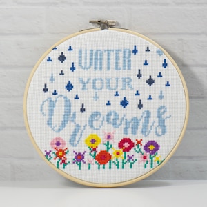 May include: A circular cross-stitch design in a wooden hoop, featuring the words "WATER YOUR DREAMS" in blue. The design includes blue droplets and a row of colourful flowers. The background is a white brick wall.