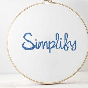 Puede incluir: Un aro de bordado blanco con la palabra "Simplify" bordada en hilo azul.