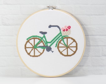 May include: Cross-stitch embroidery of a green bicycle with brown wheels and a basket of pink flowers. The bicycle is set against a white background and framed in a wooden hoop. A charming, handmade piece.