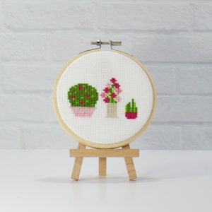 May include: A cross-stitch embroidery hoop with a design of three potted plants. The plants are a green bush, a pink flower, and a green cactus. The hoop is on a wooden easel.