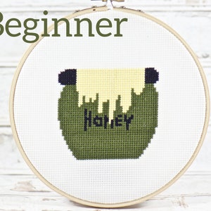 May include: A beginner cross stitch project featuring a green cauldron with a yellow liquid dripping from the top. The cauldron has the word "Honey" stitched on the front.