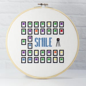 Può includere: Un cerchio da ricamo bianco con un disegno a punto croce che presenta una macchina fotografica, la parola "SMILE" in blu e una griglia di quadrati colorati.