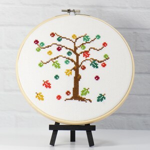 May include: A cross-stitch embroidery hoop with a colorful autumn tree design. The tree has brown branches, green and yellow leaves, and red and orange berries. The background is white.