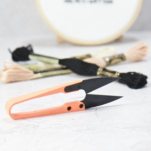 May include: A small, pink-handled embroidery scissors with black blades. The scissors are lying on a white surface with several spools of embroidery floss in the background.