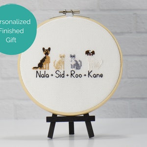 May include: A circular wooden embroidery hoop containing white fabric with a cross-stitch design of four animals, two dogs and two cats, above the embroidered black text Nala + Sid + Roo + Kane. The hoop is displayed on a small black easel.
