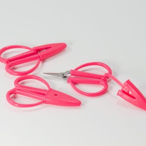 May include: Three pairs of bright pink thread snips with matching plastic protective caps. Each tool features a looped handle design and sharp metal blades. One pair is shown with the cap removed, revealing the pointed steel cutting edges.