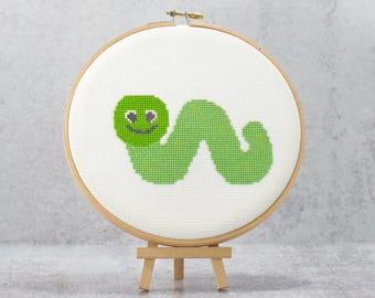 May include: A cross-stitch embroidery hoop featuring a smiling green caterpillar on a white background. The caterpillar has a green head and body, with a smiling face. The embroidery hoop is displayed on a small wooden easel.