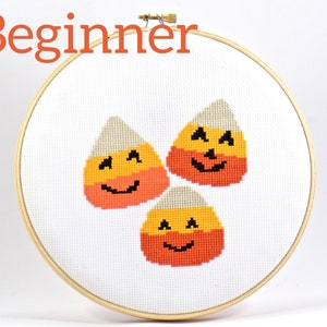Peut inclure: Trois motifs de point de croix en forme de maïs sucré orange, jaune et blanc avec des visages de citrouille sur un fond blanc dans un cercle de broderie en bois. Débutant.