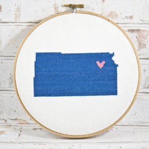 May include: A blue embroidered outline of the state of Kansas with a pink heart in the center, displayed in a round wooden embroidery hoop.