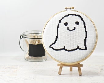 Puede incluir: Un aro de bordado blanco con un diseño de punto de cruz en blanco y negro de un fantasma sonriente. El aro está en un caballete de madera.