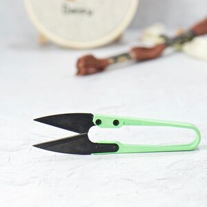 May include: A pair of thread snips featuring sharp, black metal blades and a light green plastic handle. The tool is designed for precision cutting of threads and yarn, with a spring-loaded mechanism that keeps the blades open for repeated use.