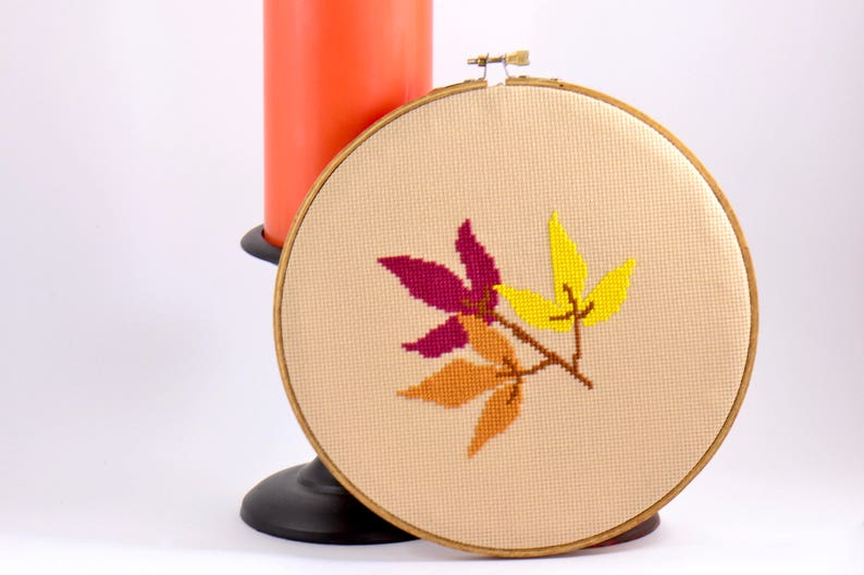 May include: A beige embroidery hoop with a cross-stitch design of three autumn leaves in shades of yellow, orange, and burgundy.
