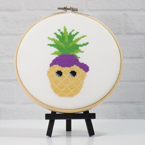 Puede incluir: Bordado de punto de cruz de una piña sonriente con la parte superior verde, sombrero morado y ojos azules. El bordado está colocado en un aro de madera y se exhibe en un pequeño caballete negro. El fondo es una pared de ladrillos blancos.