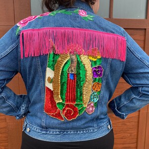 May include: A blue denim jacket with a decorative design. The back is adorned with a sequined image, pink fringe, and floral embroidery. The jacket has a button-up front and long sleeves.