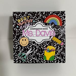 May include: A black and white composition book with a rainbow, smiley face, scissors, crayons, and pencil. The book says "Composition Book" and "Ms. Davis" in pink letters.