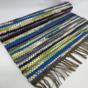 May include: A rolled-up, striped rug with a fringe. The rug features alternating stripes of blue, green, and off-white. The fringe is a mix of brown and tan. The rug is made of woven fabric.