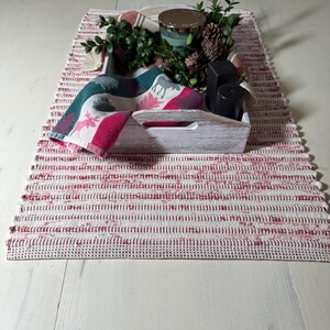 May include: A rustic table setting featuring a red and white striped runner. A wooden tray holds a candle, greenery, pine cones, and black containers. A folded napkin with a deer pattern adds a festive touch.