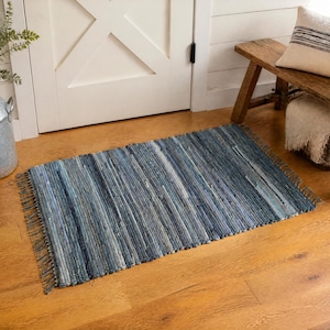 May include: A rectangular woven rag rug featuring a variegated pattern of blue, navy, and white fabric strips. The rug has a textured, multi-tonal appearance with short, frayed fringe detailing along the two shorter edges, creating a casual look.