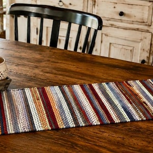 May include: A rectangular table runner featuring a multi-colored striped pattern made of woven fabric strips. The design incorporates various shades of red, blue, orange, white, and grey, creating a textured, linear appearance across the entire surface.