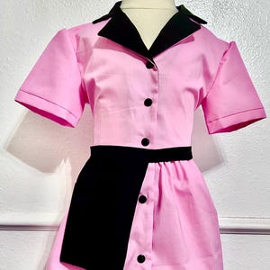 May include: A pink dress with a black collar, short sleeves, and a black asymmetrical panel. The dress has black buttons down the front and a black belt. The dress is displayed on a mannequin.