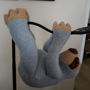 May include: A plush sloth toy in shades of gray, tan, and brown, hanging upside down. The sloth has a light gray body, tan hands and feet, and a brown face with black eyes. The toy is hanging from a black bar.