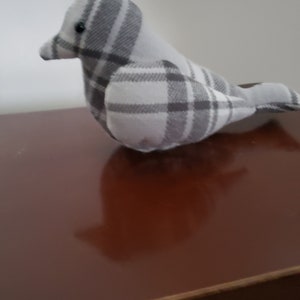 May include: A gray and white plaid bird plush toy. The bird is sitting on a brown wooden surface.