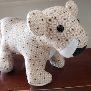 May include: A plush toy lion with a patterned fabric body in beige, brown, and black. The lion has large black button eyes, white felt around the mouth, and a black nose. The toy is standing on four legs.