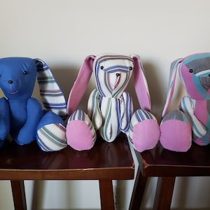 May include: Three handmade striped fabric bunny plushies. The bunnies are sitting on a wooden surface. The bunny on the left is blue with white and gray stripes. The bunny in the middle is white with pink, gray, and blue stripes. The bunny on the right is pink with gray, white, and blue stripes.
