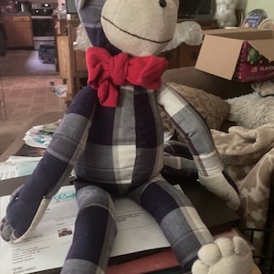 May include: A handmade stuffed monkey doll with a plaid pattern in navy blue, gray, and white. The monkey has a cream-colored face, ears, hands, and feet, and a bright red bow tie. The doll is sitting on a table.
