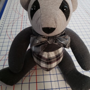 May include: A gray and black teddy bear with a plaid bow tie. The bear is sitting on a white gridded surface.