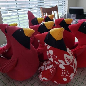 May include: A group of eight red cardinal bird plush toys. The birds are all sitting in a row, and they have black eyes, yellow beaks, and white accents on their chests. Some of the birds have a floral pattern on their chests.