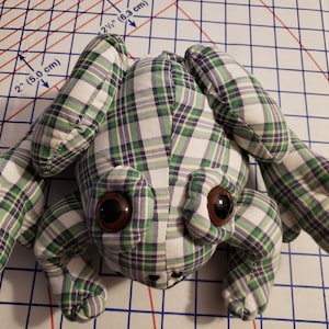May include: A handmade frog plushie in a green, white, and purple plaid pattern. The frog has large brown eyes and is lying on a grid-lined surface. The plushie has four limbs and a small, stitched mouth.