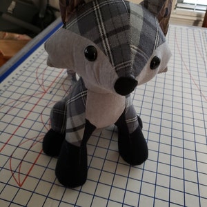 May include: A gray and black plaid fox plush toy with black button eyes. The toy is sitting on a white gridded surface.