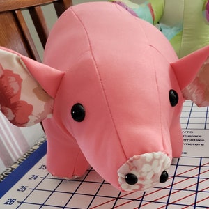 May include: A pink pig stuffed animal with floral fabric ears. The pig has black button eyes and a white floral snout.
