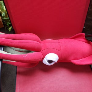 May include: A red plush squid with a white eye and green tentacles. The squid is lying on a red chair.
