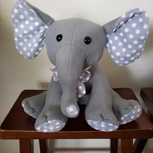 May include: A gray plush elephant with large ears and a trunk. The elephant's ears, feet, and bow are patterned with white polka dots. The elephant has black button eyes and is sitting on a wooden stool.