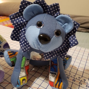 May include: Blue and white fabric lion stuffed animal with a white polka dot mane. The lion is sitting on a blue and white gridded surface. The lion's legs are made of comic book fabric.