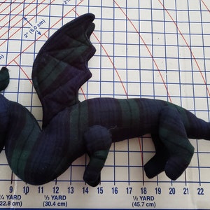 May include: A black and green plaid dragon plush toy measuring approximately 20 inches long. The dragon is lying on its side with its wings spread out.