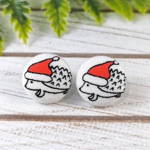 May include: A pair of white fabric button earrings with a black and white illustration of a hedgehog wearing a red Santa hat.