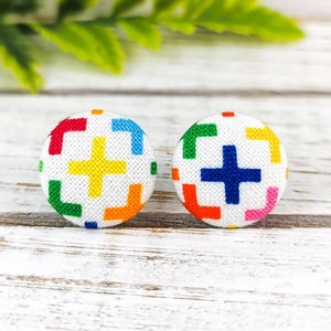 May include: A pair of colourful fabric button earrings with a white background and a plus sign design. The buttons are round and have a geometric pattern of squares and rectangles in shades of red, orange, yellow, green, blue, and pink.