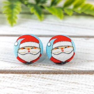 May include: A pair of round, blue and white fabric earrings with a Santa Claus design. The Santa Claus has a red hat, white beard, and a red suit.