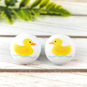 May include: A pair of white button earrings featuring a yellow rubber ducky design. The ducky is floating in blue water.